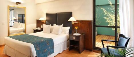 A hotel room with a large bed, a nightstand, a chair, and a view of greenery through a sliding door.