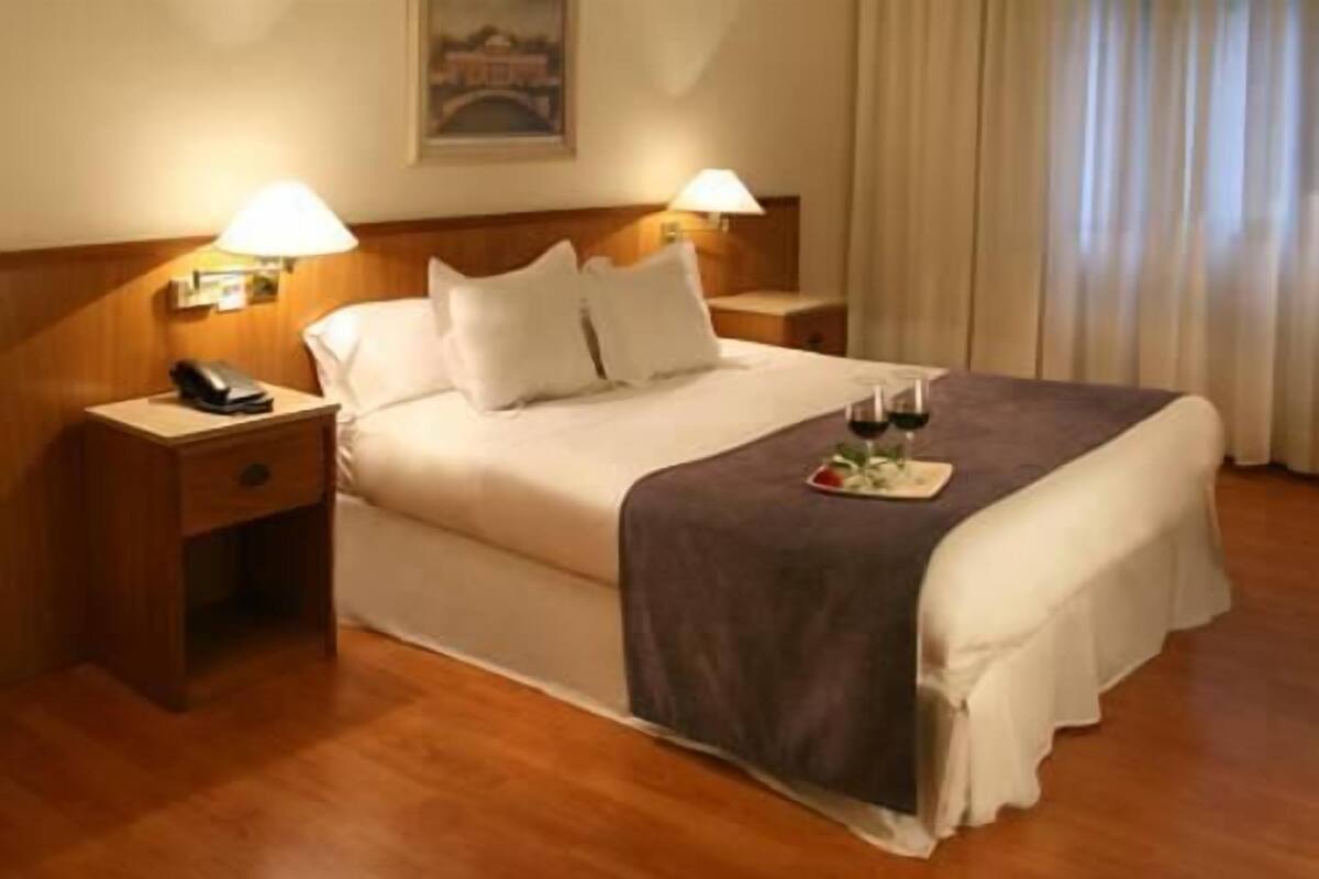 A hotel room with a bed, bedside lamps, a nightstand, and a tray with a glass of wine.