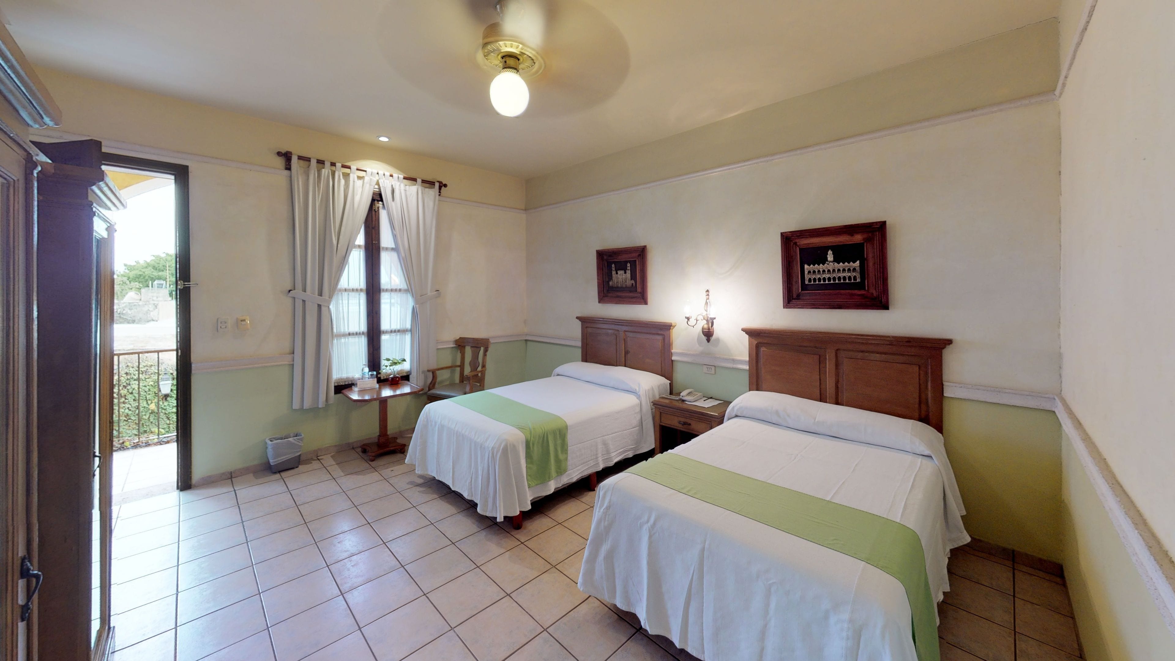 Una habitación de hotel con dos camas, una ventana con cortinas, una mesita y una lámpara de pared.