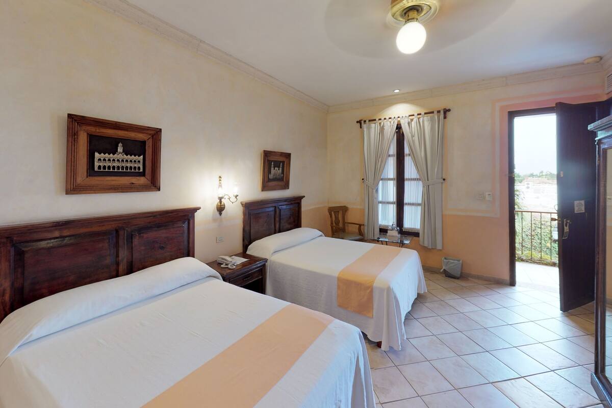 A hotel room with two beds, a wooden headboard, a nightstand with a phone, a window with curtains, and a door leading to a balcony.