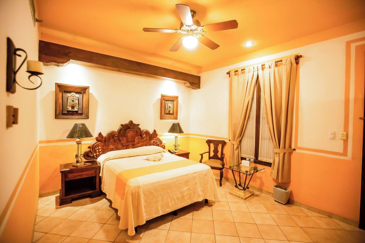A hotel room with a large bed, a ceiling fan, two bedside tables with lamps, a chair, and a window with curtains.