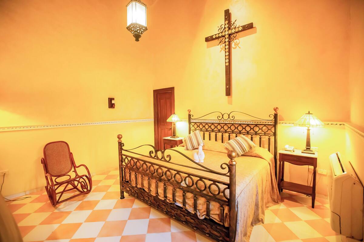 A room with a checkered floor, a bed with a metal frame, two bedside lamps, a chair, and a cross on the wall.