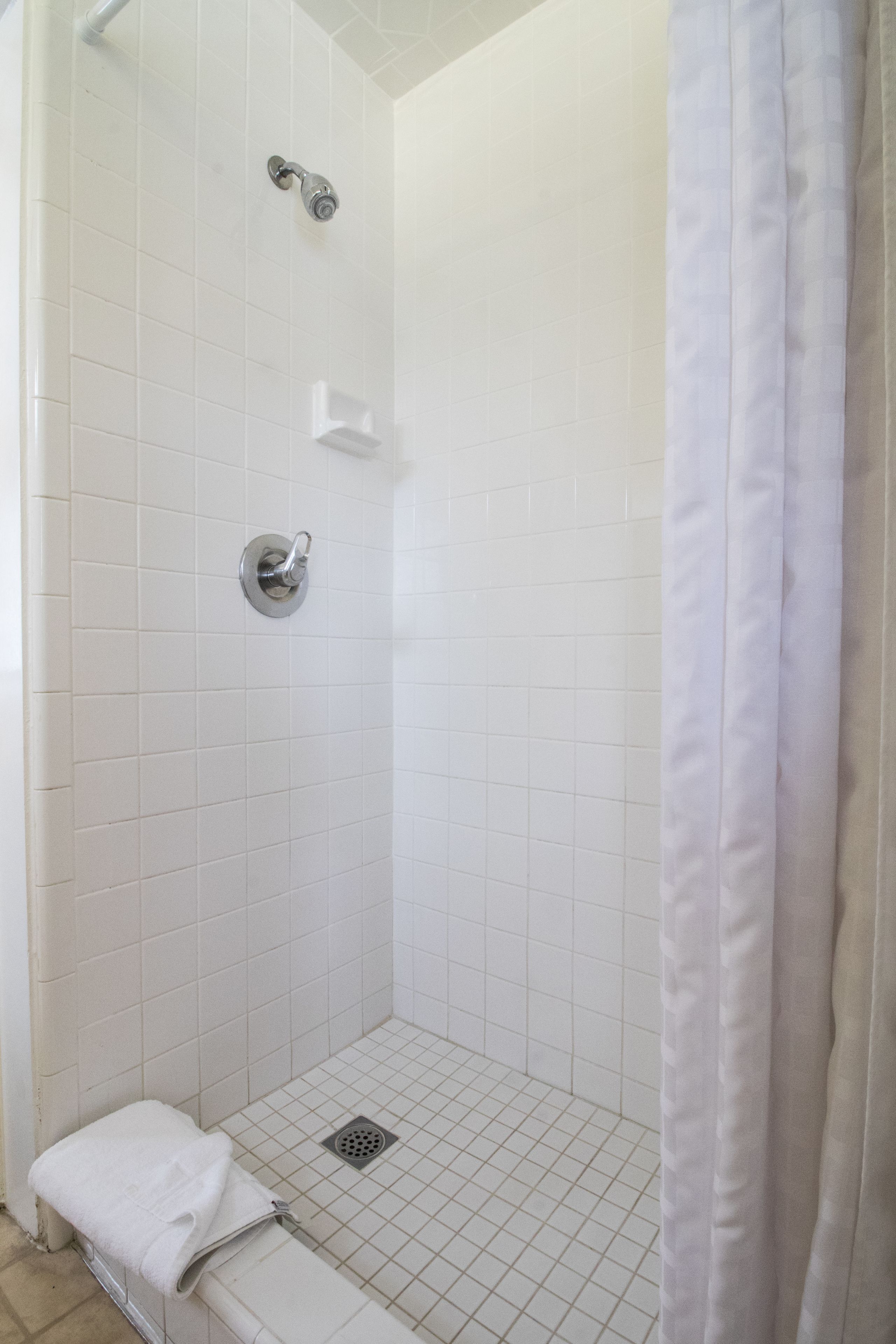 A white tiled shower with a showerhead, a towel, and a drain.