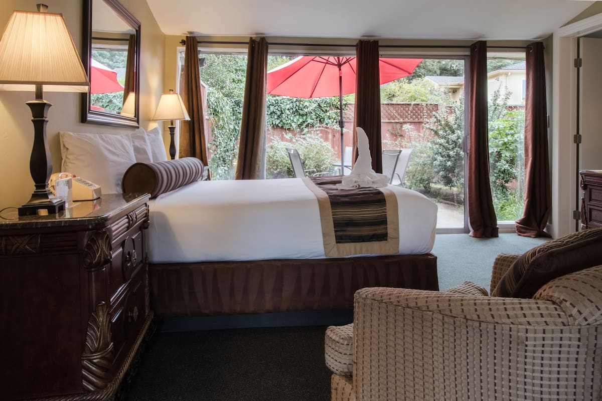 A hotel room with a large bed, a wooden dresser, a chair, and a view of the garden through the window.