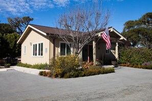 A single-story house with a front porch, an American flag, and a small garden with various plants and flowers.