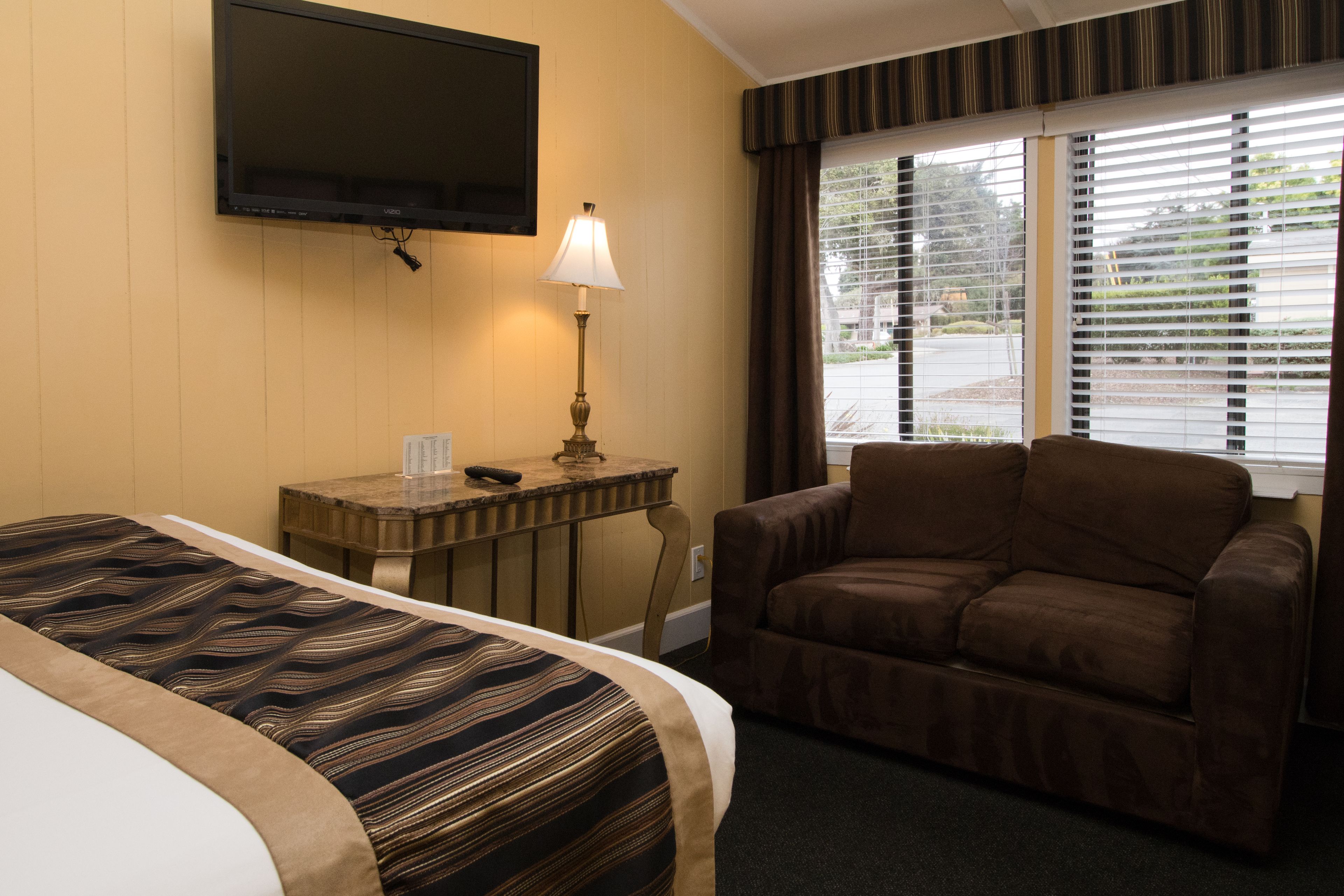 Une chambre d’hôtel avec un lit, un bureau doté d’une lampe, un fauteuil brun et un téléviseur fixé au mur.