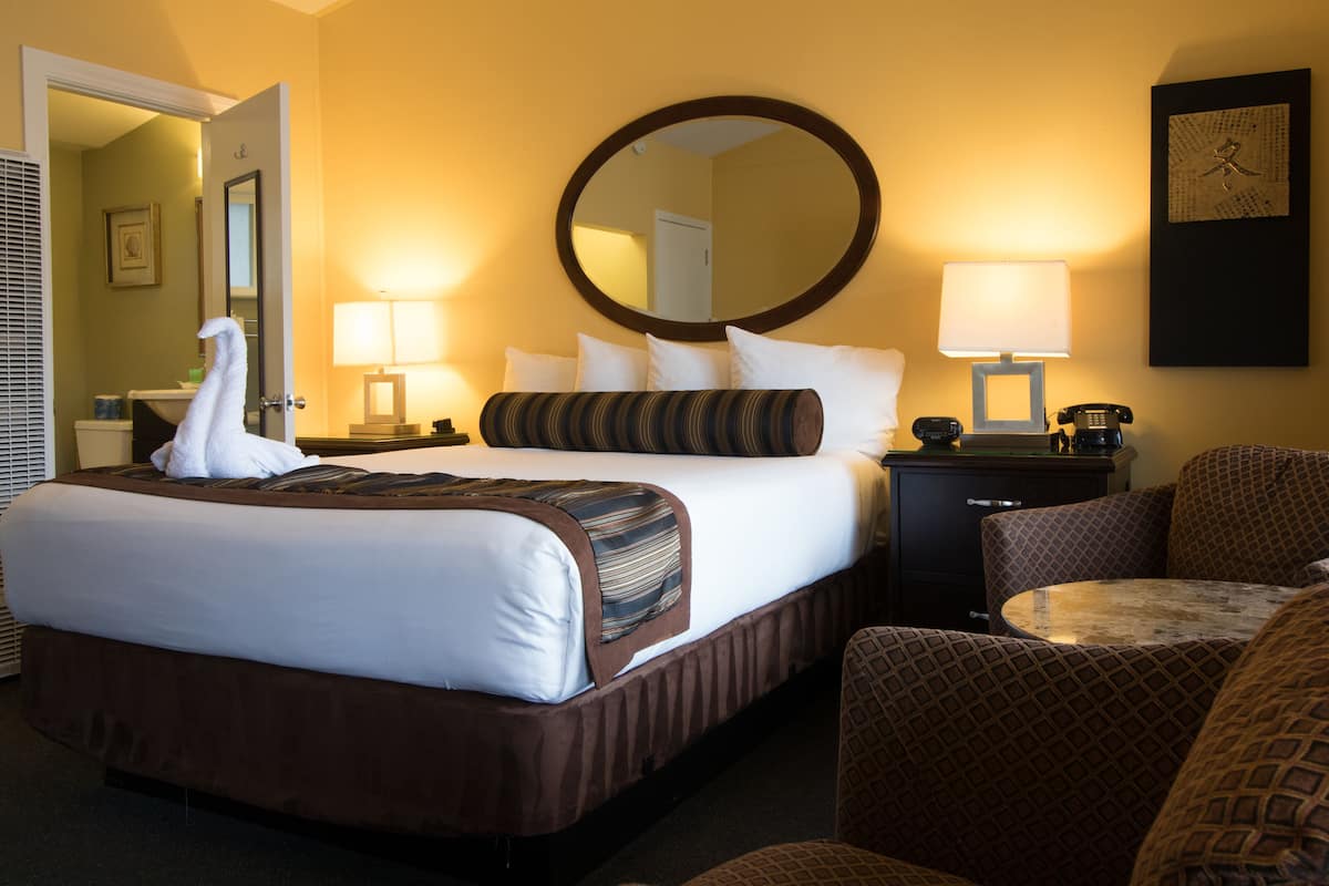 A hotel room with a large bed, two bedside tables with lamps, a round mirror, a patterned armchair, and a small round table.