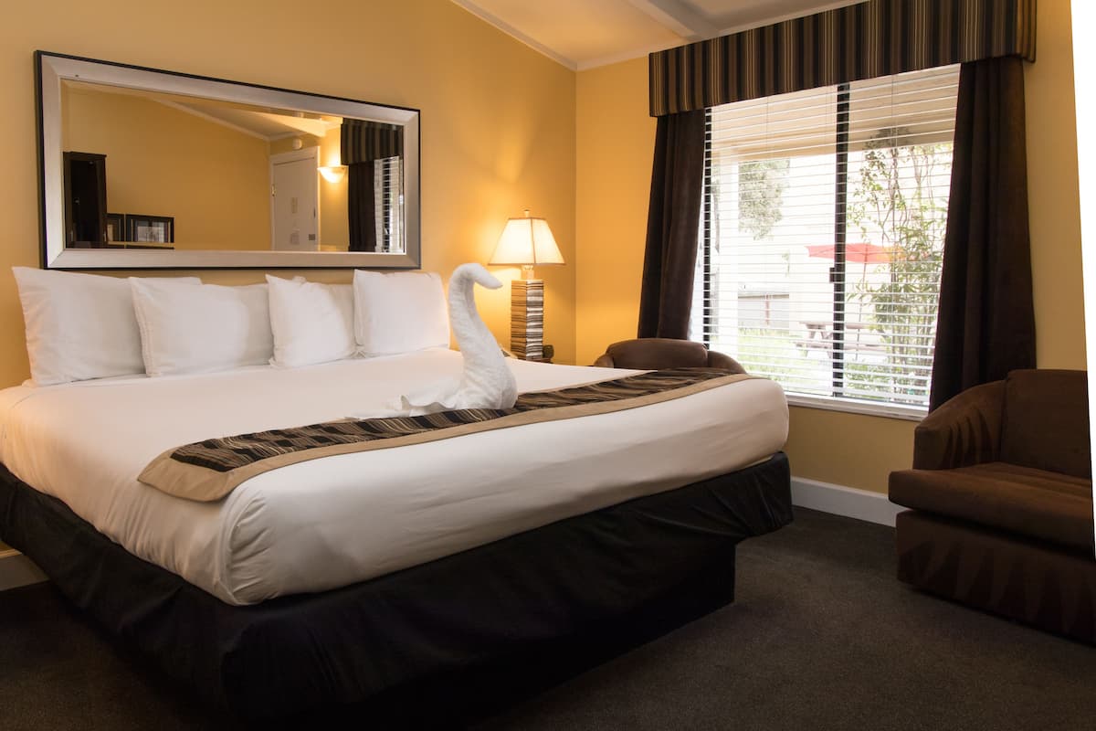 A hotel room with a large bed, a mirror, a lamp, a brown armchair, and a window with blinds.
