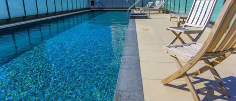 Una piscina en la azotea con agua azul cristalina, sillones de madera y barandilla de vidrio.