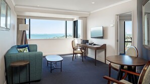 A hotel room with a sofa, a small table, a desk with a chair, a TV, and a large window offering a view of the sea. - The Spencer Suites By Castle (Auckland)
