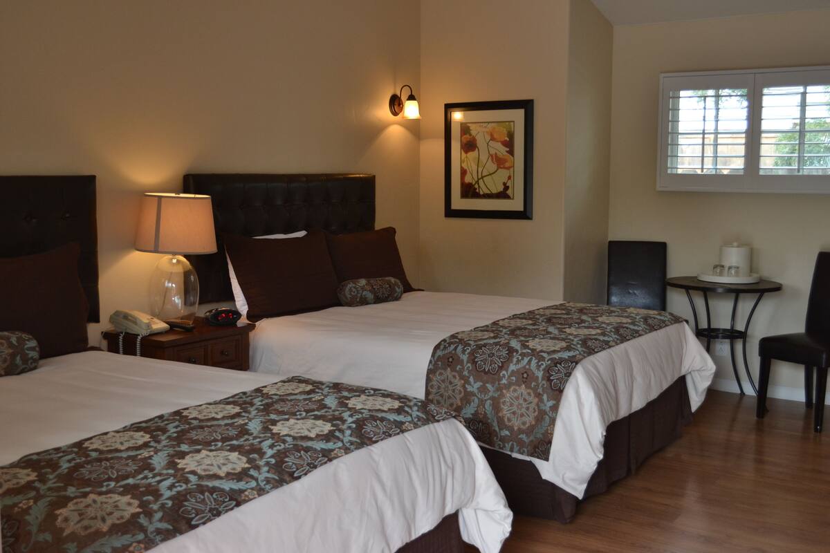 Quarto de hotel com duas camas, uma mesinha de cabeceira com abajur, uma cadeira e uma pequena mesa com xícara e pires.