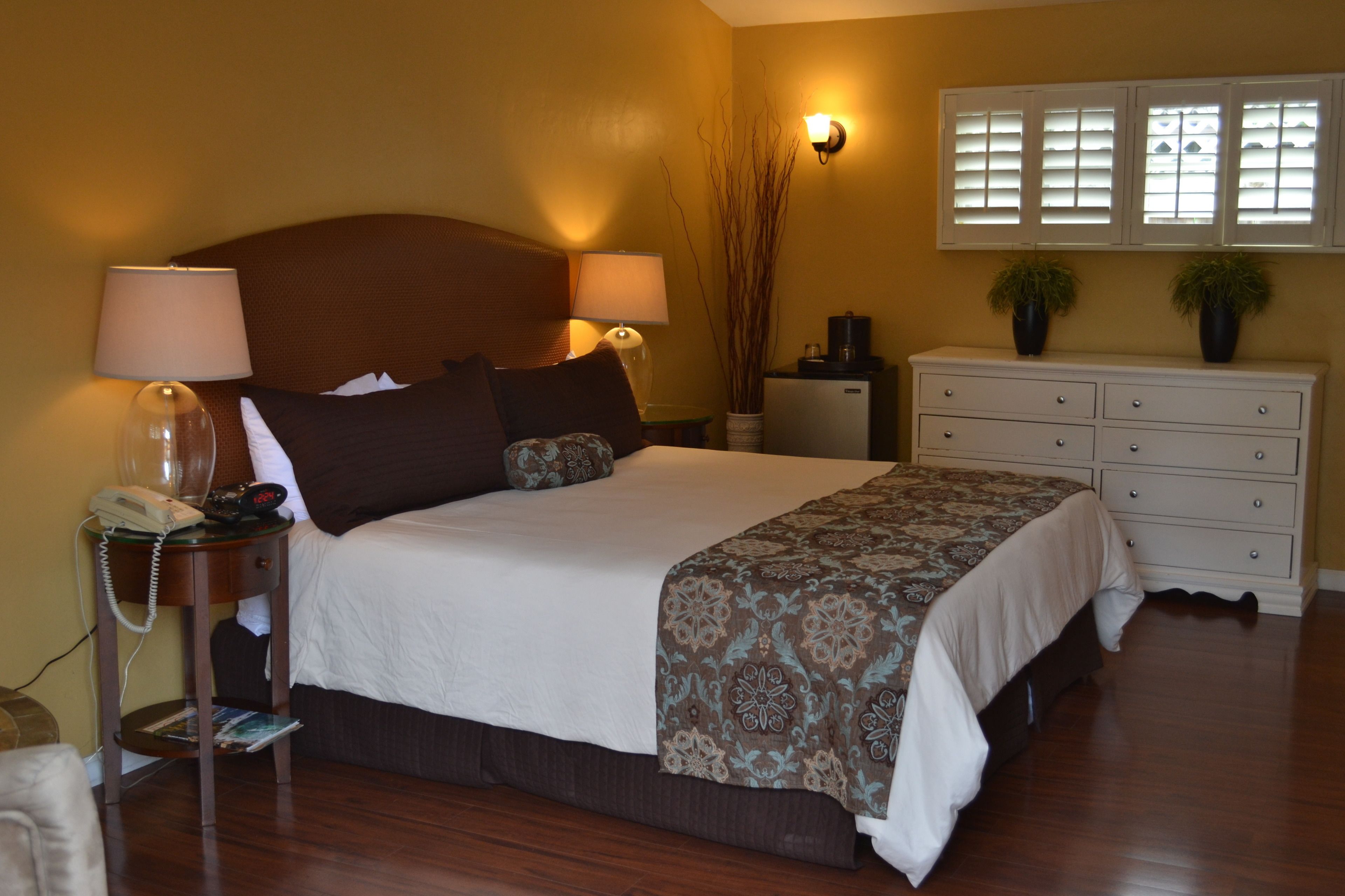 Habitación de hotel con una cama grande, lámparas de noche, mesita de noche con teléfono, cómoda y ventana con persianas.