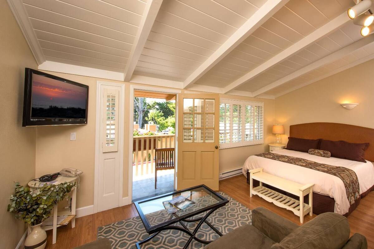 Quarto com cama, TV, mesa de centro de vidro, um banco e porta que dá para uma varanda.