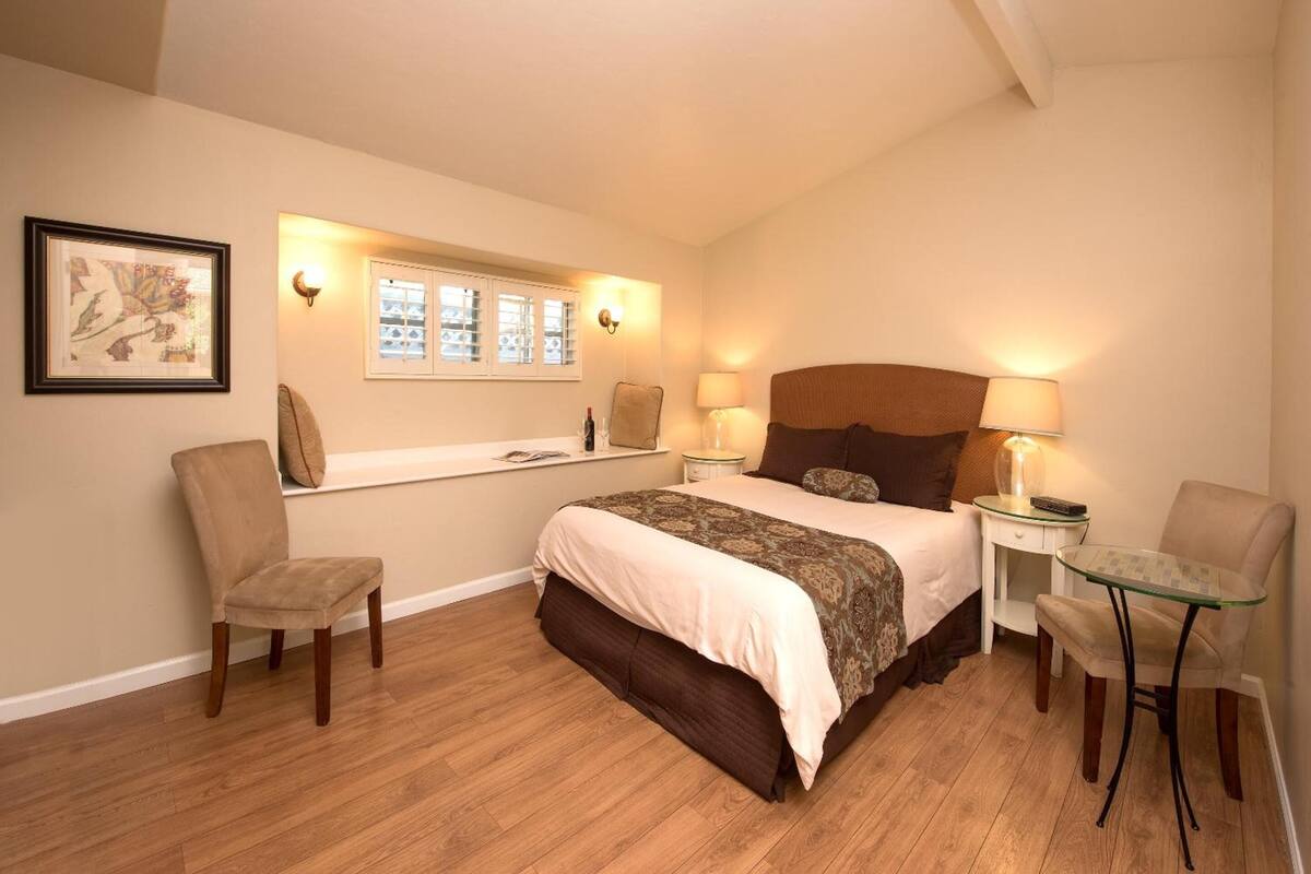 Quarto com cama, duas cadeiras, uma mesinha, mesa de cabeceira, janela e luminárias fixadas na parede.