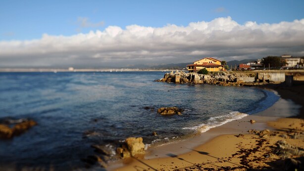 A coastal scene with a building on a rocky outcrop, a sandy beach, and a clear sky. - Sunset Inn (Pacific Grove)