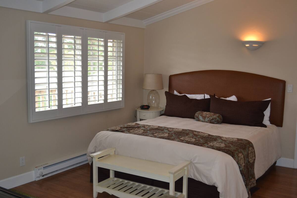 Quarto com cama grande, mesa de cabeceira, luminária e janela com persianas.
