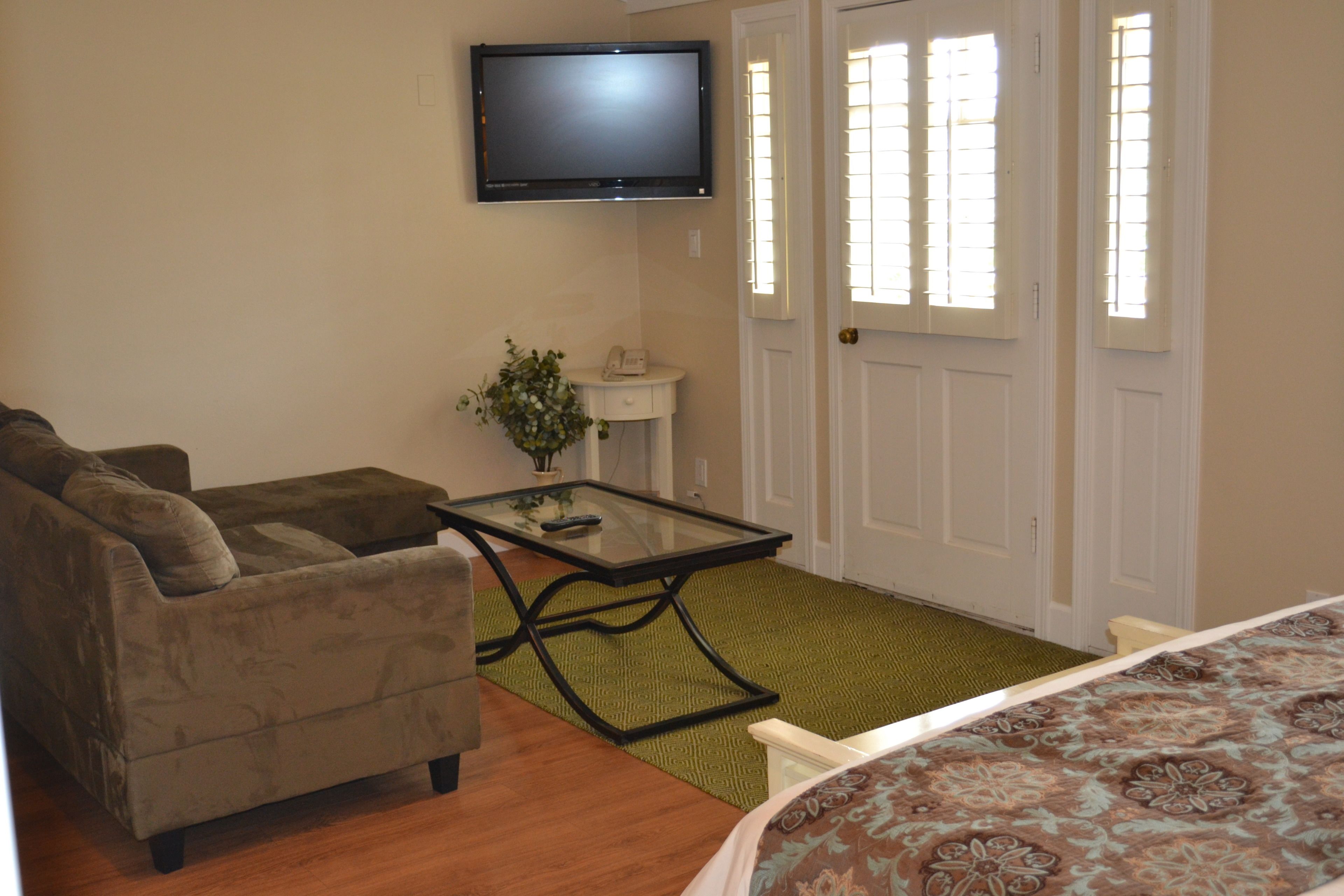 A room with a sofa, a glass-top coffee table, a flat-screen TV mounted on the wall, a small side table with a lamp, and a door with windows.