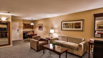 Une chambre d’hôtel comprenant un canapé, deux fauteuils, une table basse, une lampe, un téléviseur, un coin cuisine et un grand tableau encadré au mur.