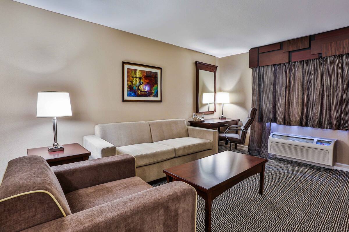 Une chambre d’hôtel avec un canapé beige, une table basse en bois, un bureau avec une lampe, un grand miroir et une fenêtre avec des rideaux.