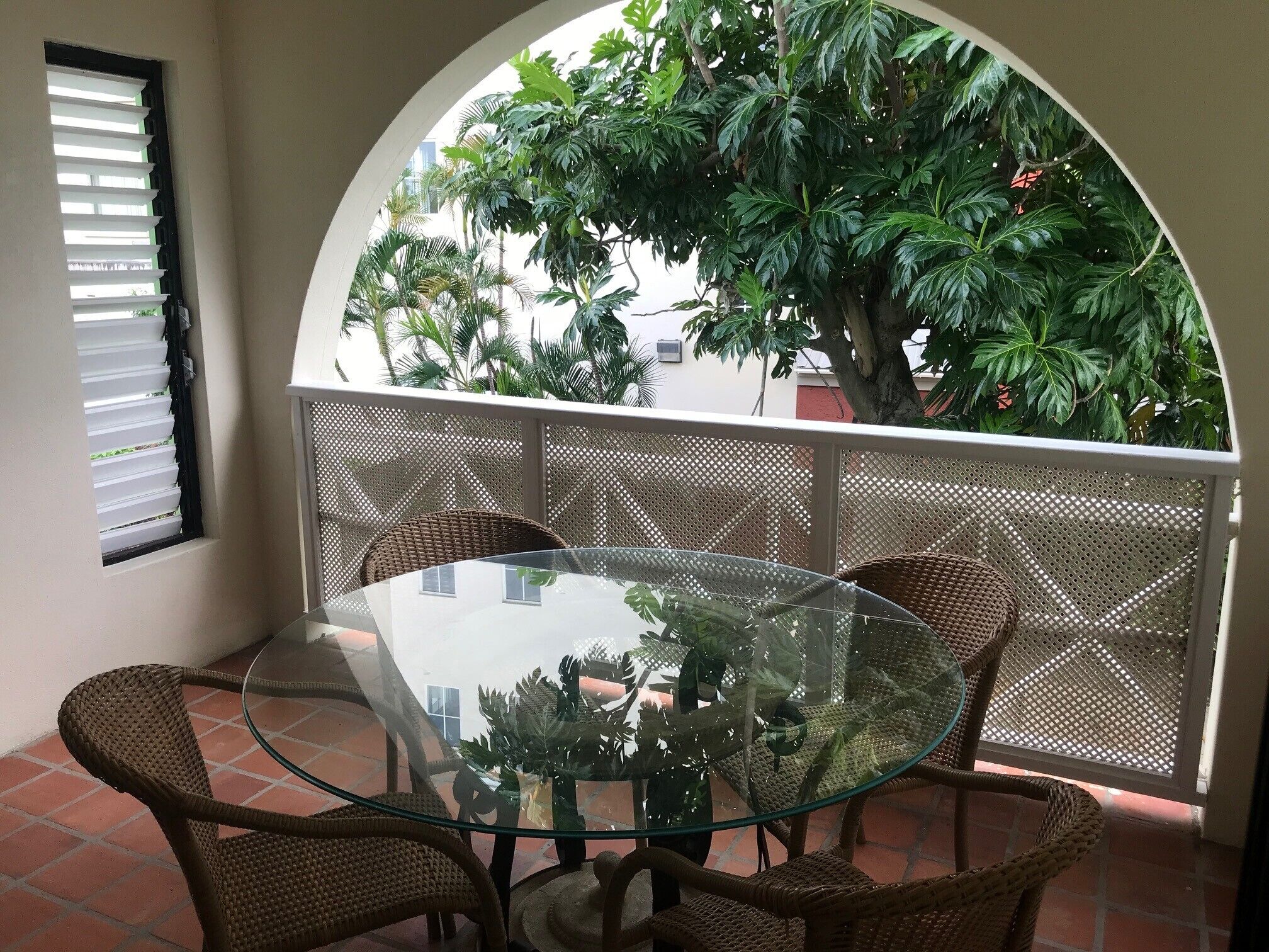 A glass-top table with wicker chairs, an arched window, and a view of a tree and building.