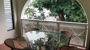 Una mesa con cubierta de vidrio, sillas de mimbre, un ventanal arqueado y vistas a un árbol y un edificio.