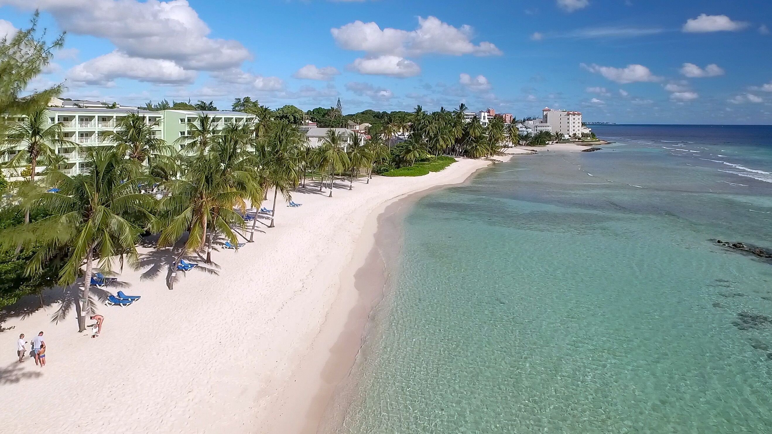 A beach with clear turquoise water, white sand, and palm trees. There are buildings with balconies and a hotel in the background.