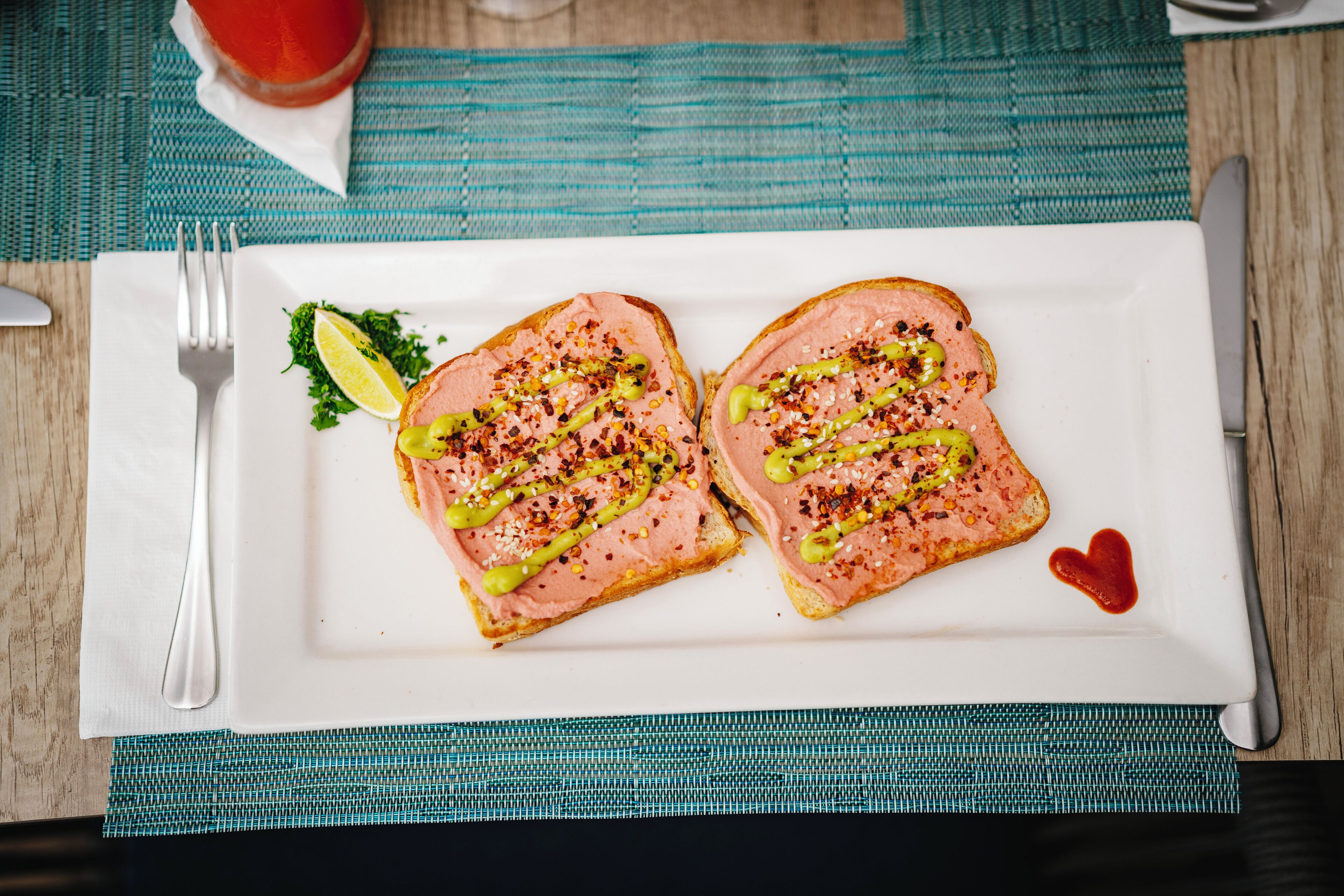 Een wit rechthoekig bord met twee sneetjes toast, belegd met roze spread, groene saus en zaden, vergezeld van een partje citroen en een kleine decoratie in de vorm van een hart.