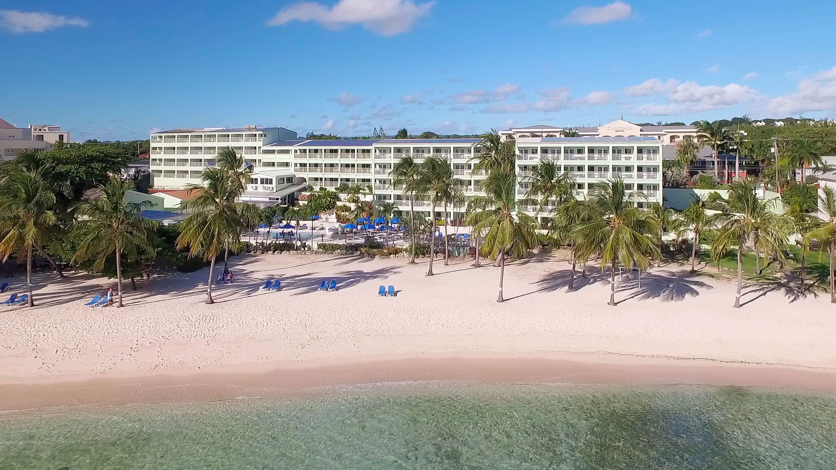 A beachfront hotel with multiple floors, a pool area with umbrellas, and palm trees.