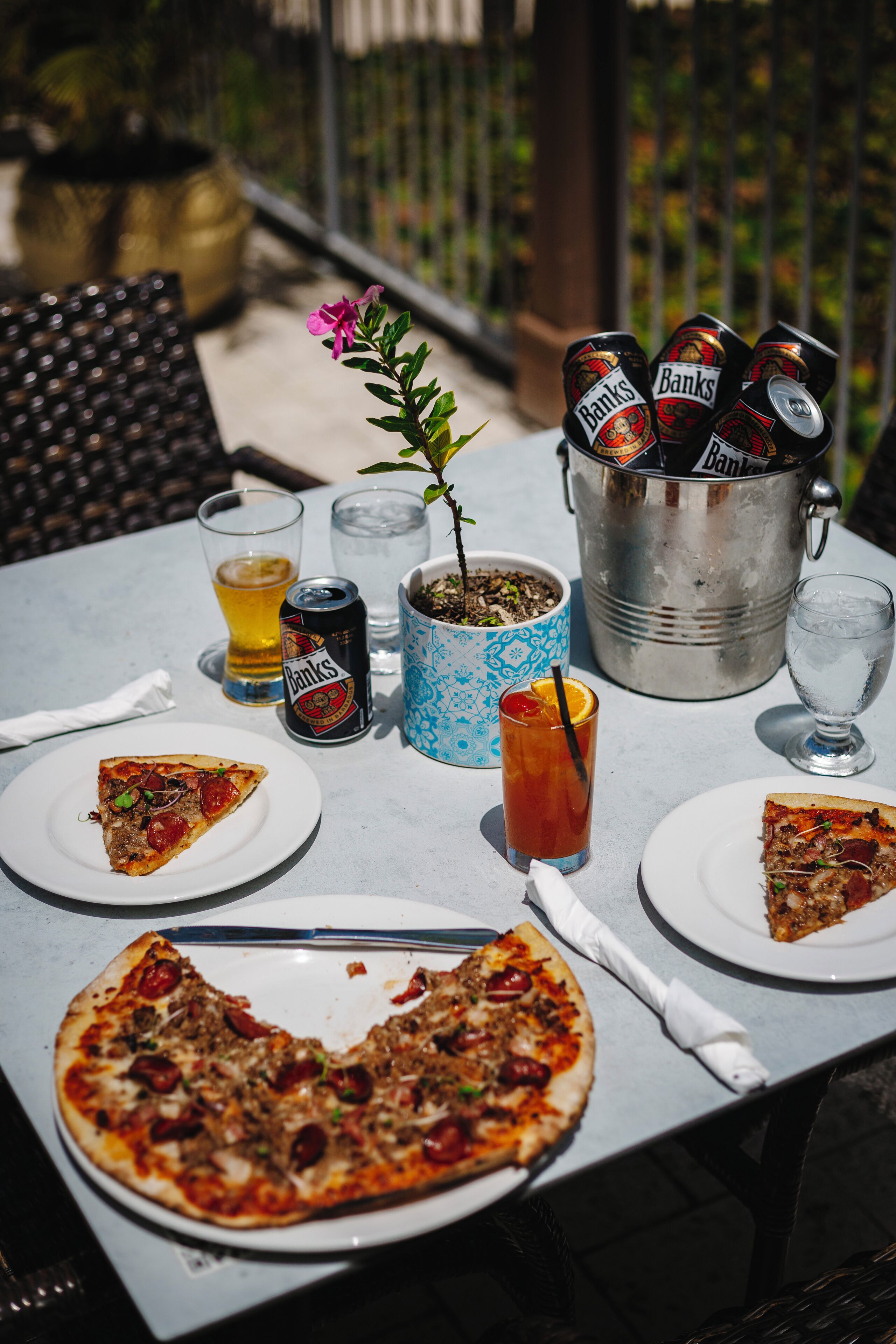 A table with pizza slices, a drink, a can of Banks beer, and a bucket of ice.