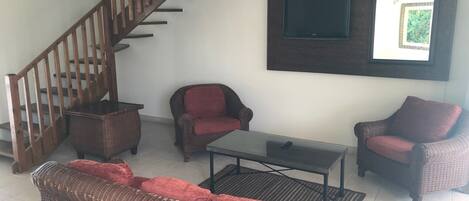 Una sala de estar con escalera de madera, muebles de mimbre, una mesa de centro de vidrio y un televisor montado.