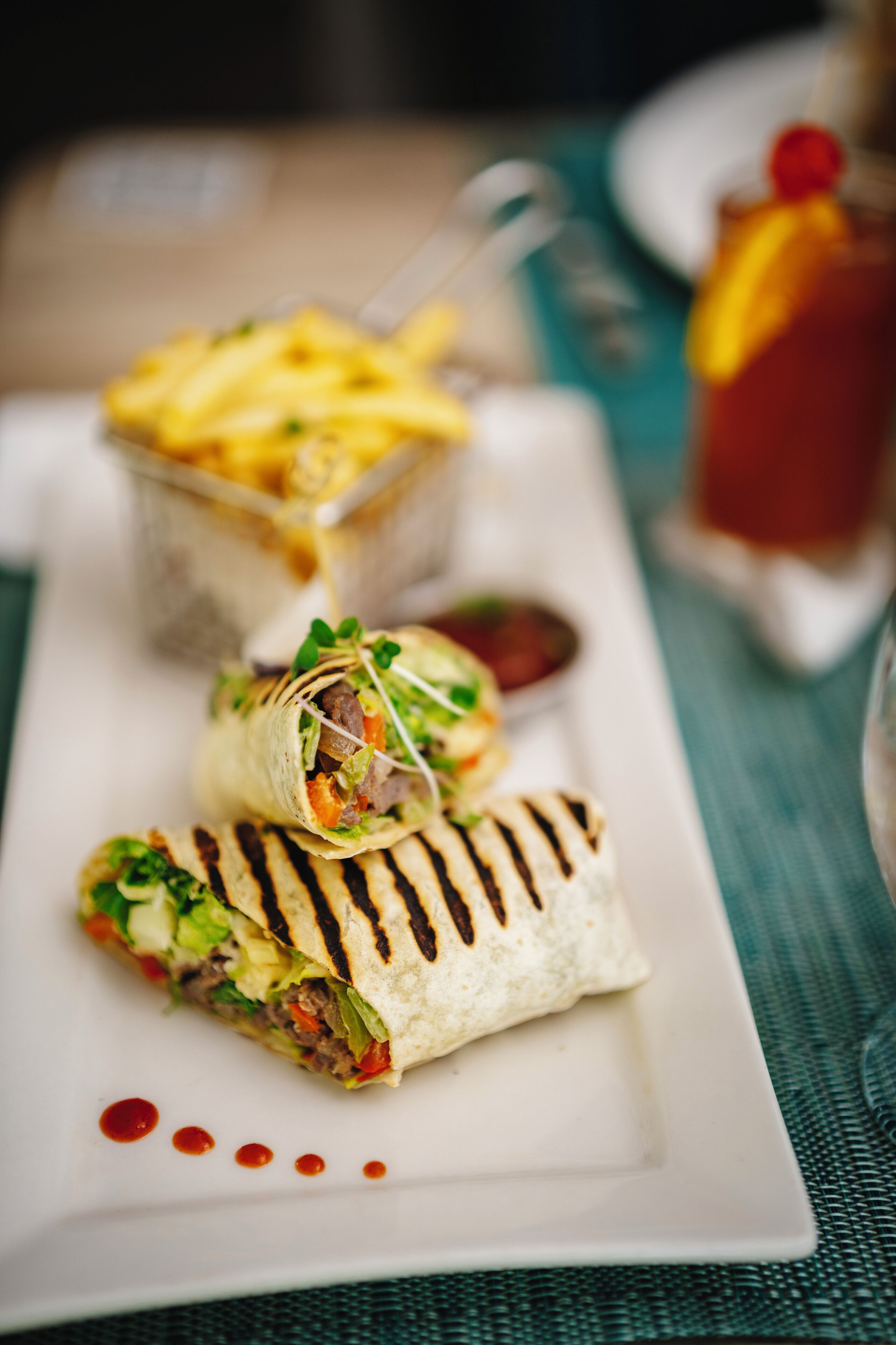 A plate with a wrap and fries, and a drink in the background.