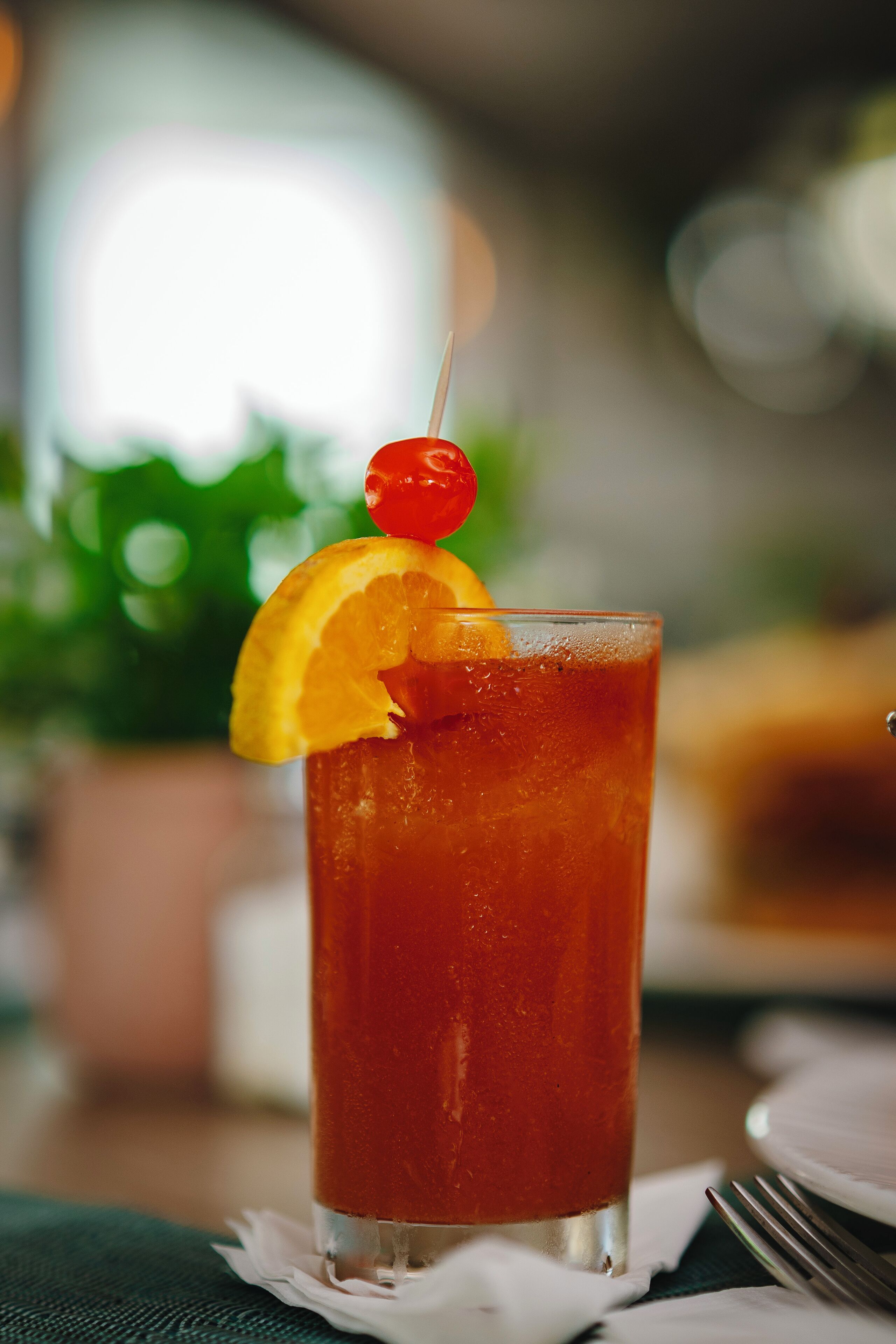 A tall glass of iced tea with a cherry and orange slice garnish, placed on a table with a fork and napkin.