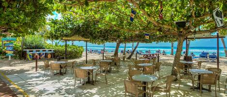Un café al aire libre con mesas y sillas bajo un árbol, con vista a una playa con gente y barcos.