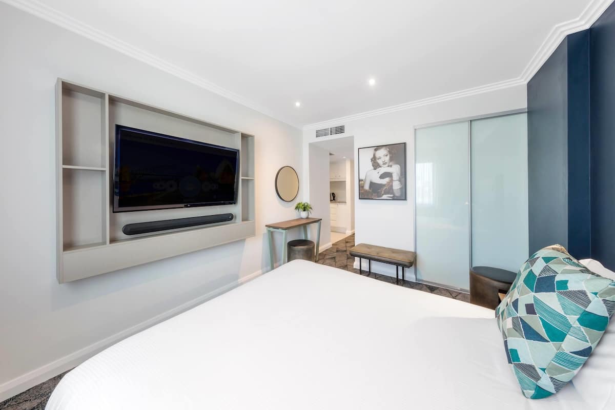 Quarto de hotel moderno com uma cama grande, televisão de tela plana na parede, uma mesinha com uma planta e um guarda-roupa com portas deslizantes.