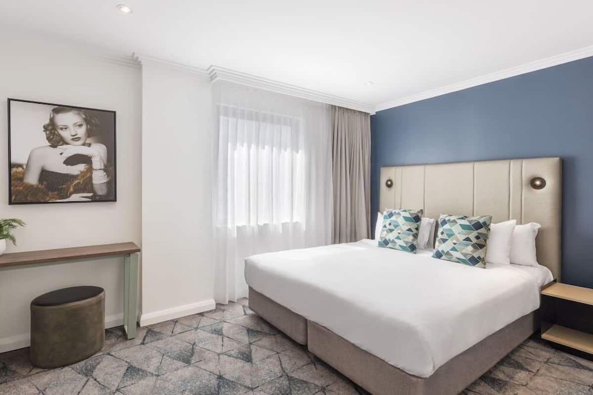 Quarto de hotel moderno com uma cama grande, cabeceira com almofadas decorativas, mesa de cabeceira e um quadro na parede.