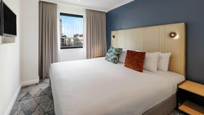 Habitación de hotel con una cama grande, un televisor montado en la pared y una ventana con cortinas.