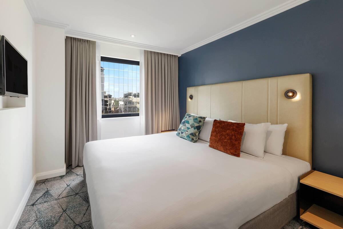 Quarto de hotel com uma cama grande, televisão fixada na parede e uma janela com cortinas.