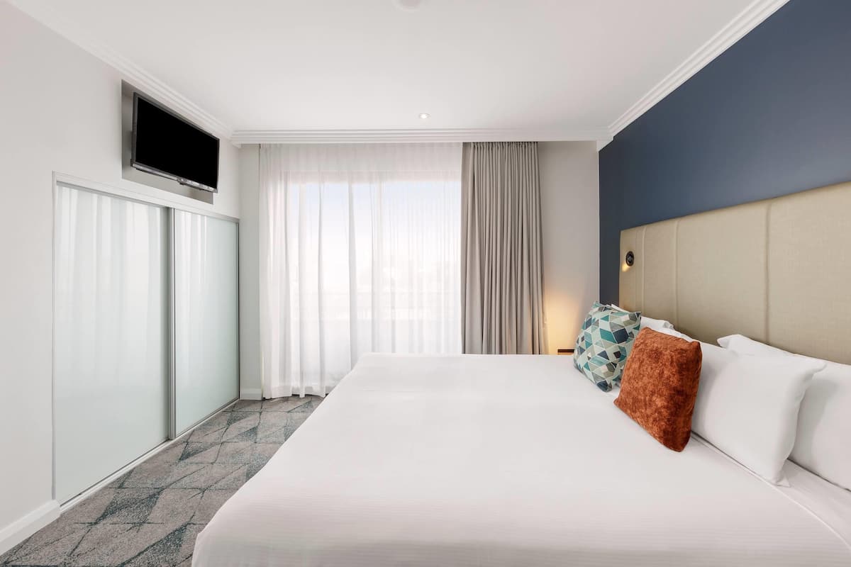 Quarto de hotel com uma cama grande, televisão de tela plana na parede, janela com cortinas translúcidas e cabeceira com luminária.