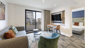 Una habitación de hotel con sofá, un sillón verde, una mesita, televisión y cama.