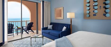 Habitación de hotel con cama, sofá, mesa de centro y balcón con sillas y vista al océano.