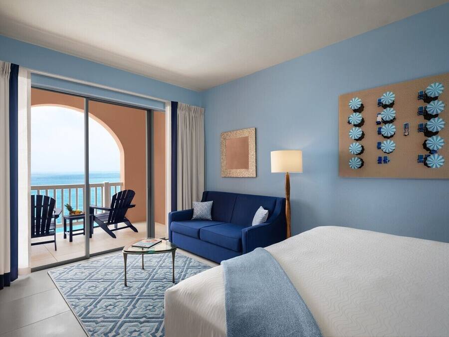Een hotelkamer met een bed, een bank, een salontafel en een balkon met stoelen en uitzicht op de oceaan.