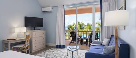 A hotel room with a bed, a desk with a TV, a sofa, a balcony with chairs, and a view of the ocean.