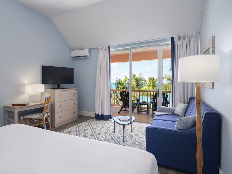 A hotel room with a bed, a desk with a TV, a sofa, a balcony with chairs, and a view of the ocean.