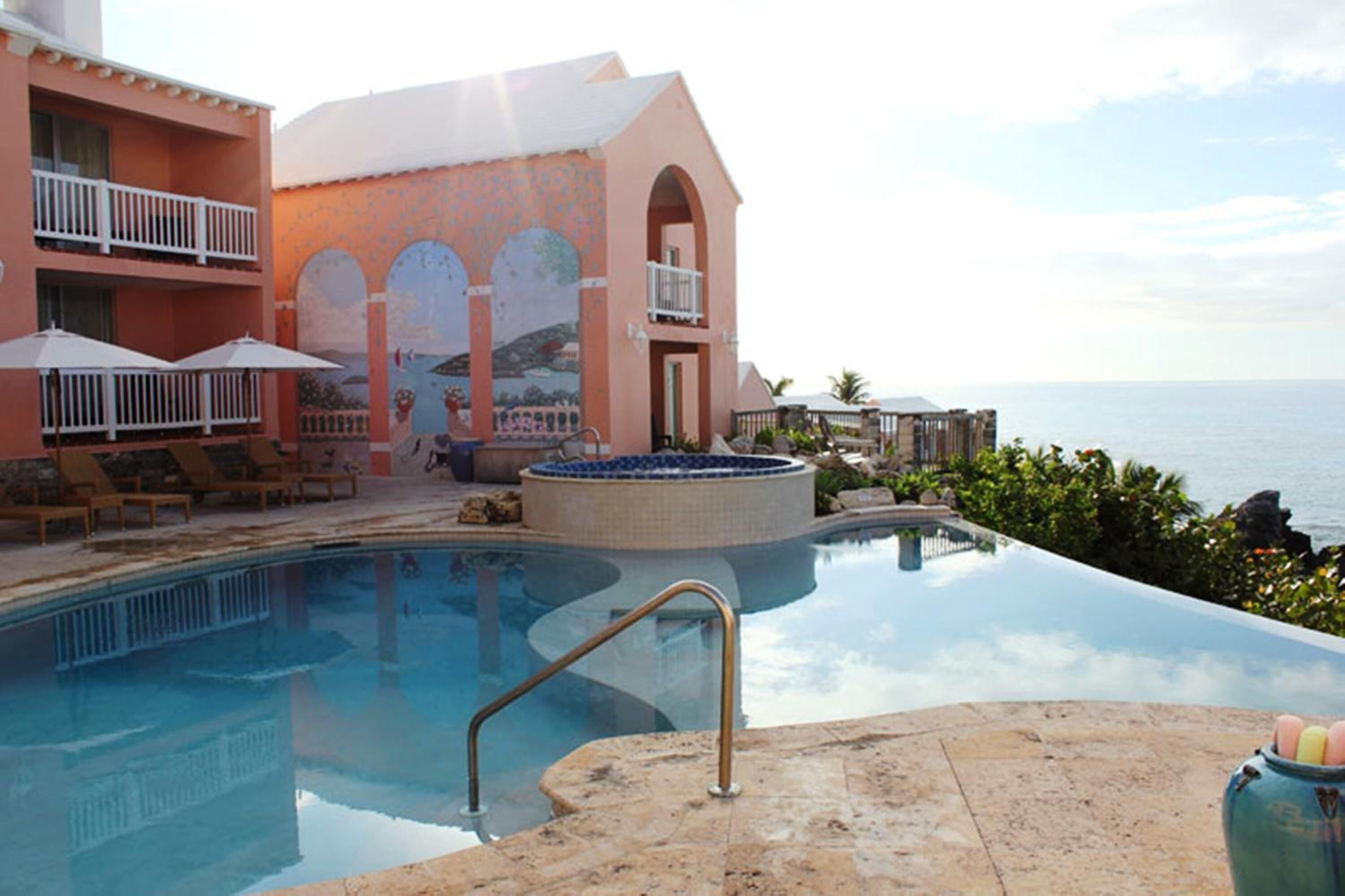 A pool area with lounge chairs and umbrellas, a hot tub, and a view of the ocean.