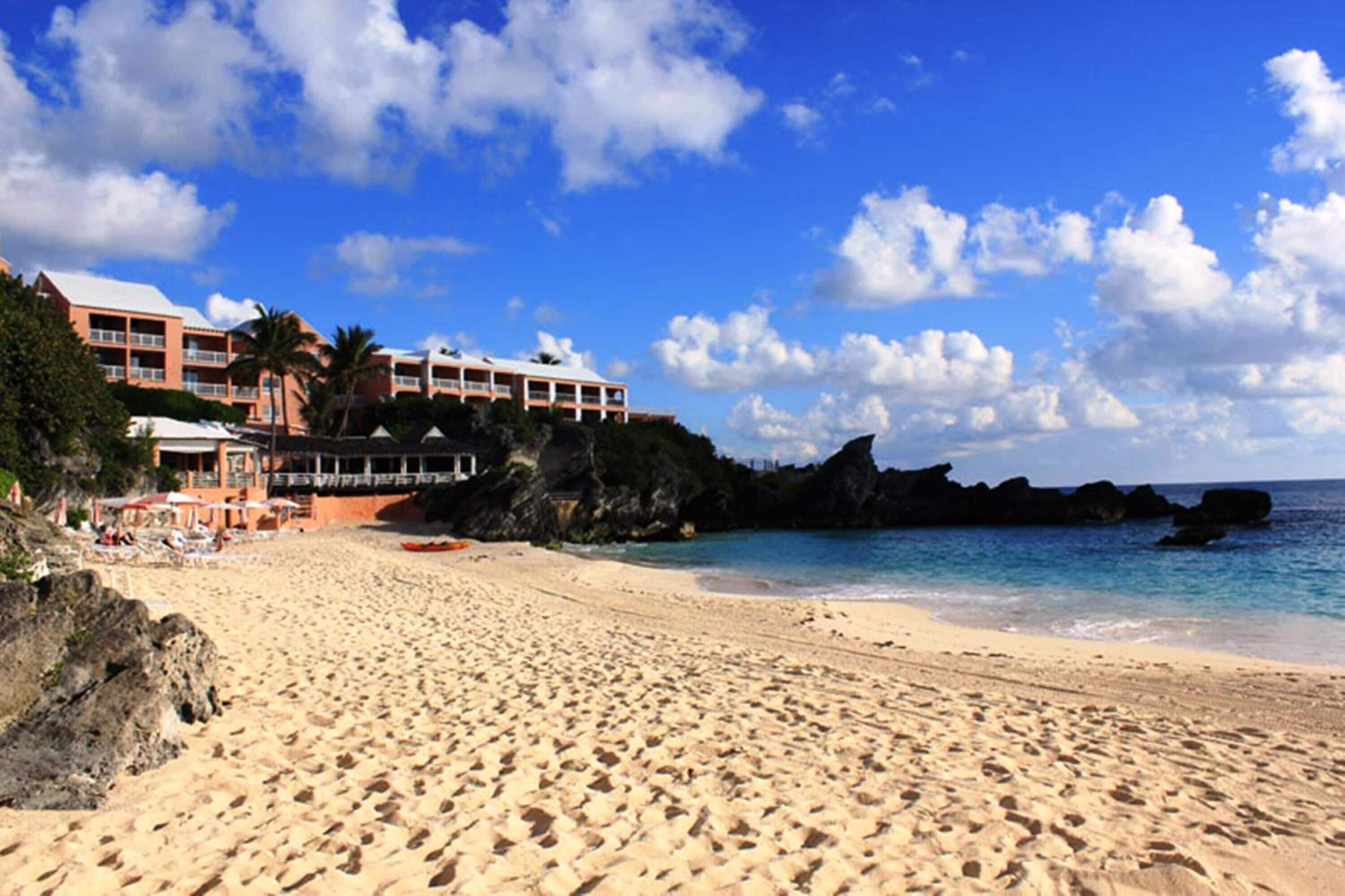 A beach with a hotel in the background, featuring a sandy shore, clear blue sky, and rocky outcrops.