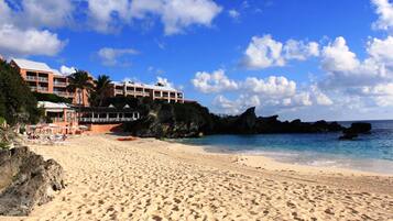 A beach with a hotel in the background, featuring a sandy shore, clear blue sky, and rocky outcrops.