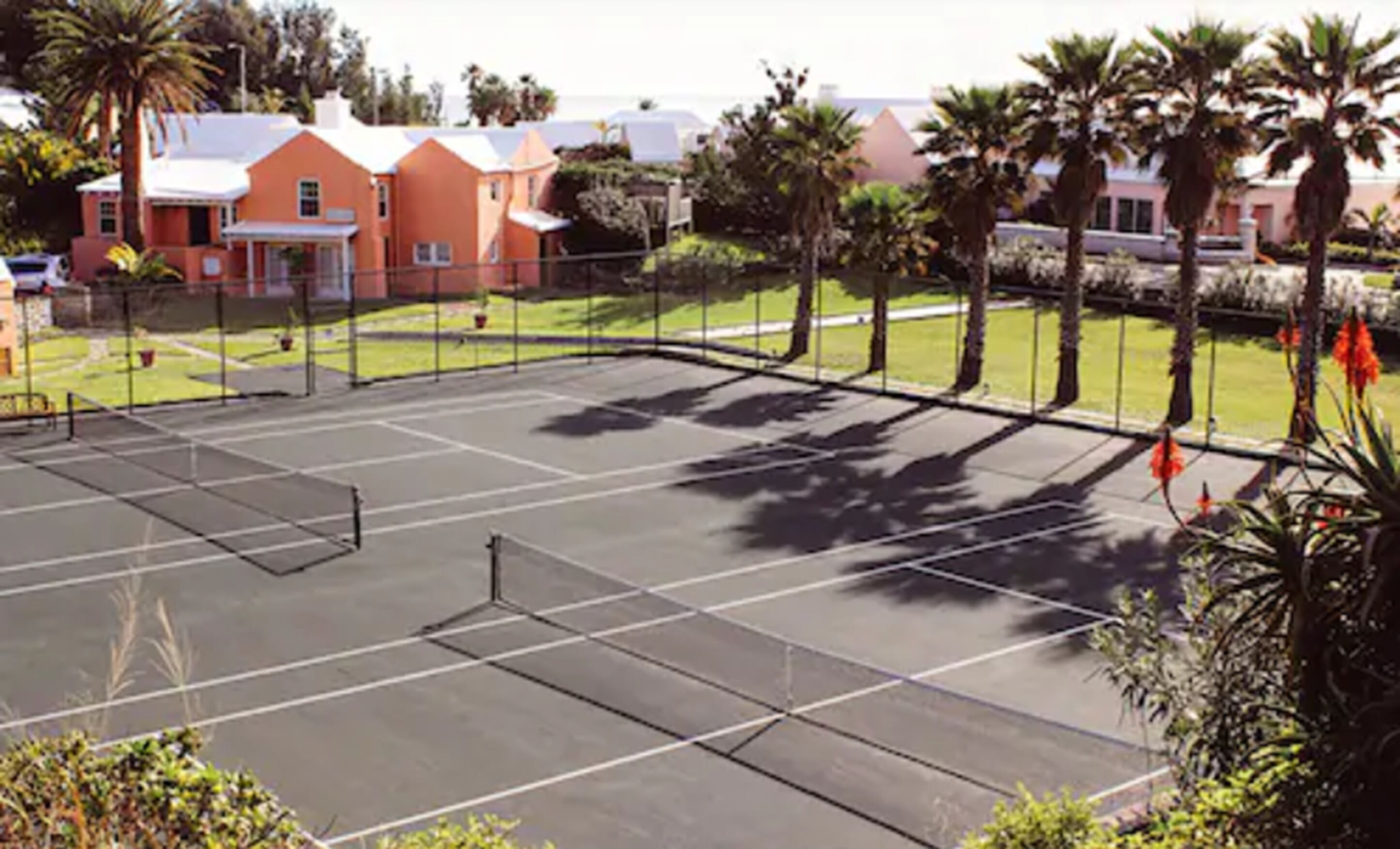 Un court de tennis avec un filet, entouré de palmiers et bordé d’un bâtiment au toit rouge.