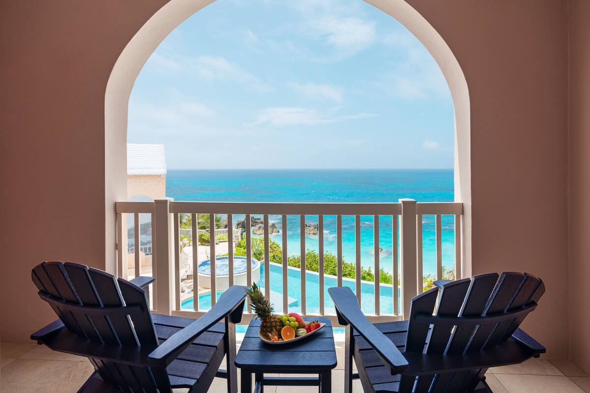 A balcony with two Adirondack chairs, a small table, and a fruit bowl. The view includes a pool and the ocean.