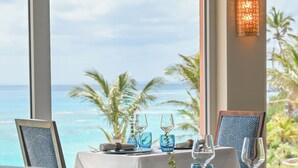 A dining table set with blue glassware and a framed picture, overlooking a beach with palm trees.