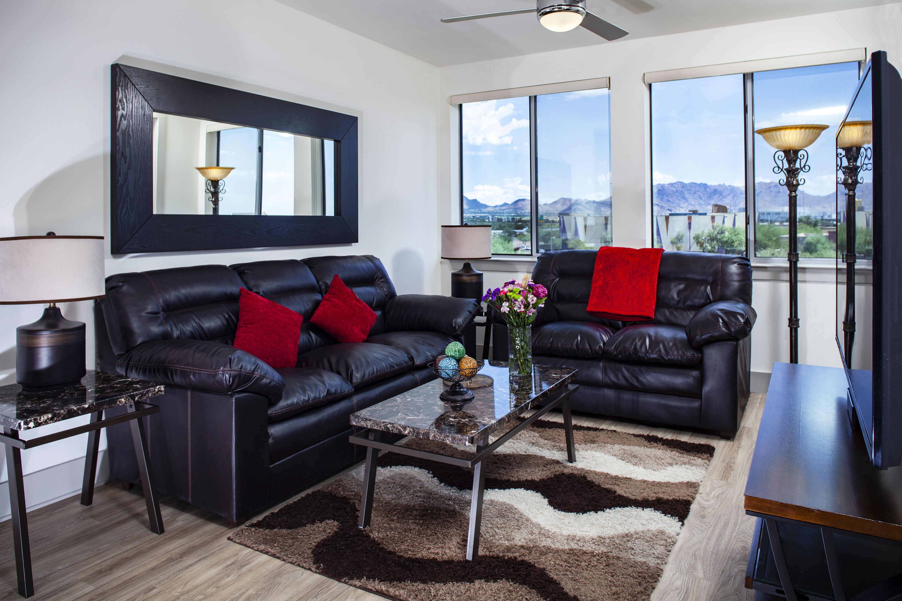 A modern living room with a black leather sofa, a glass coffee table, and a large mirror above the sofa. There are two windows providing a view of the mountains.